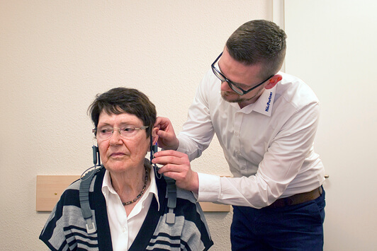 Ein Hörakustiker passt einer älteren Frau ein Hörgerät zur Hörtest-Beratung an.